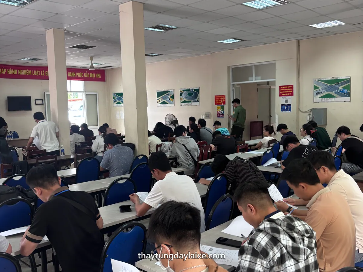 Học viên ngồi tại bàn trong hội trường, cầm giấy tờ trên tay; trên tường treo các bảng sơ đồ tình huống giao thông