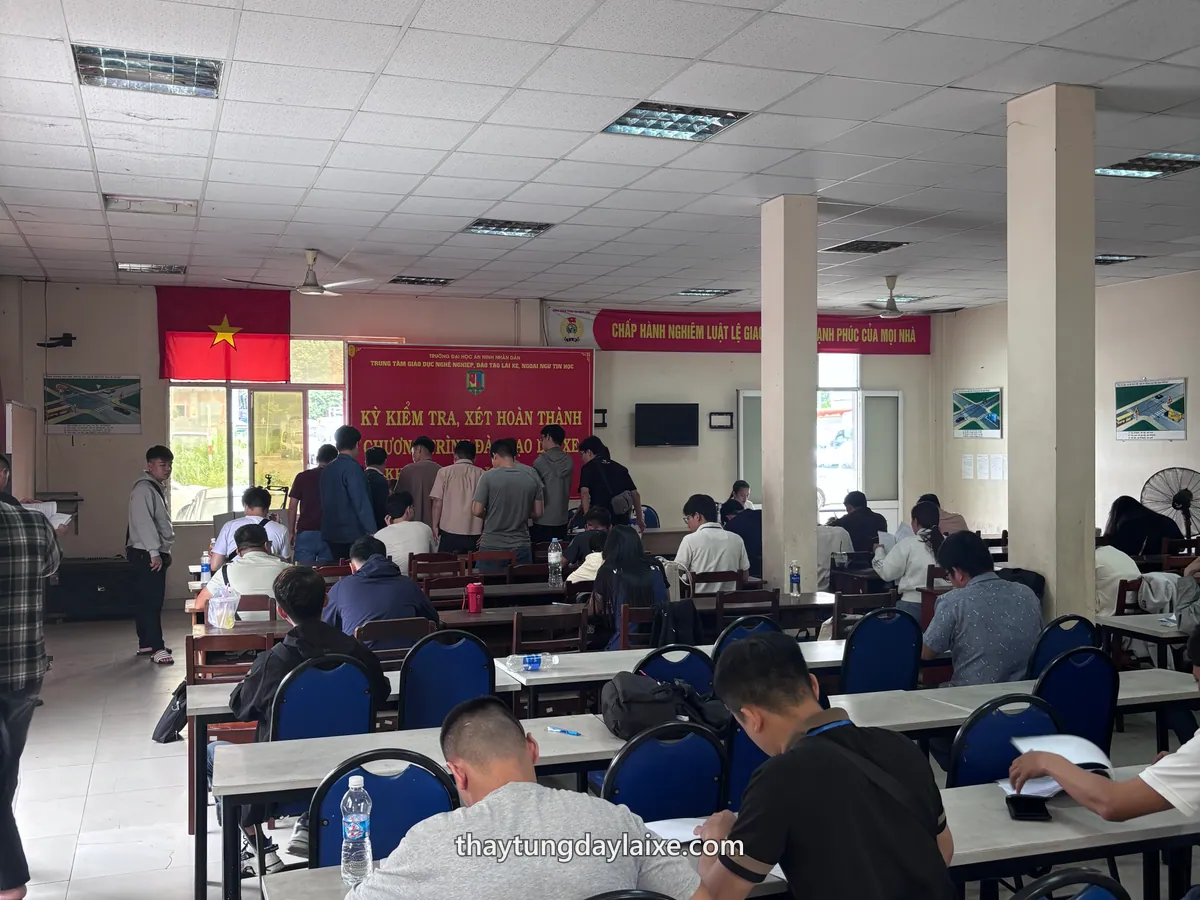 Hội trường đông học viên ngồi tại bàn, phía trước là phông đỏ của kỳ kiểm tra hoàn thành chương trình đào tạo lái xe và cờ Tổ quốc