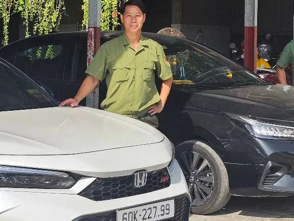 Thầy dạy lái xe mặc đồng phục xanh đứng cạnh xe ô tô, tay đặt trên nắp capo